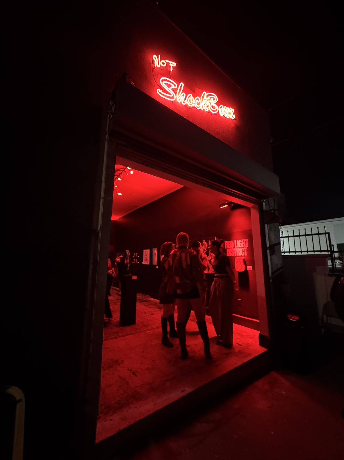 Picture of Not Shockboxx art gallery from outside with neon light lit up and inside lit in red for the show Red Light District